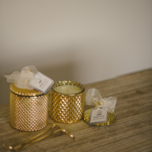 Two gold candles in cut glass vessels with diamond embossing, one large and one small, both with white wax toppers, displayed on a wooden surface.