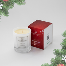 A clear candle with a red label 'Winter Season' from The Wax Company, placed next to its red packaging box with 'The Wax Company' branding on it, surrounded by artificial greenery and red berries.