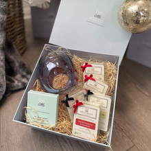A gift box open to reveal a pearl grey wax burner, four wax melts, and additional complimentary wax melt and tea lights, all arranged on a bed of decorative straw. The gift box is adorned with red ribbon bows.