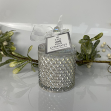 Decorative candle in a textured glass holder with a 'The Little Wax Company' tag, surrounded by greenery.