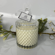 Decorative candle with a textured glass lid and 'The Wax Company' tag, surrounded by greenery.