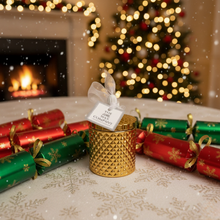 Gold candle holder with a white label in front of Christmas crackers and a decorated tree.
