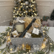 Gift basket with candles and a bottle of olive oil in front of a Christmas tree.