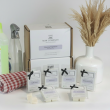 A collection of wax melt cubes from The Wax Company, displayed in front of a box with cleaning fragrances. The cubes are labeled with different scent names and are presented with a vase of dried plants and cleaning supplies in the background.