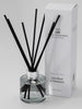 A clear glass reed diffuser with black reeds and a white label, alongside its packaging box with the brand name and fragrance details.